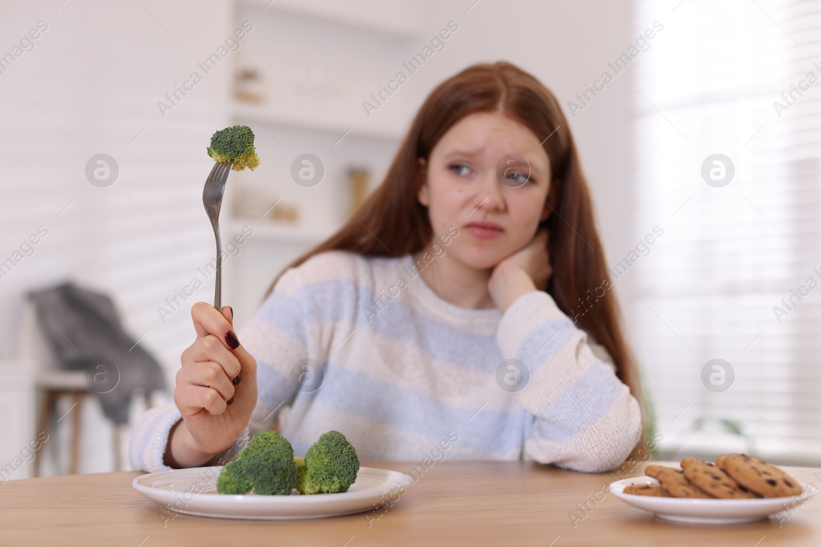 Photo of Sad teenage girl with broccoli and cookies at wooden table, selective focus. Eating disorder