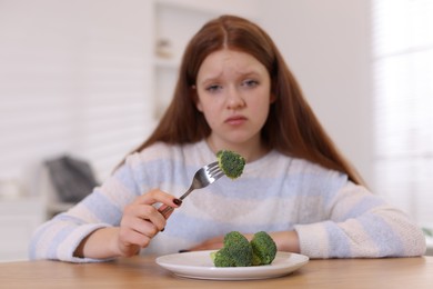 Photo of Sad teenage girl with broccoli at wooden table, selective focus. Eating disorder