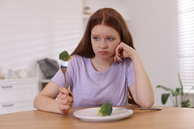 Sad teenage girl with broccoli at wooden table. Eating disorder