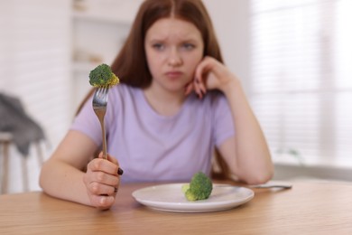 Sad teenage girl with broccoli at wooden table, selective focus. Eating disorder