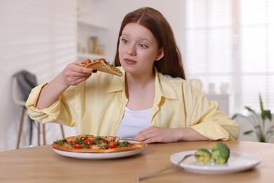 Photo of Sad teenage girl with pizza and broccoli at wooden table indoors. Eating disorder