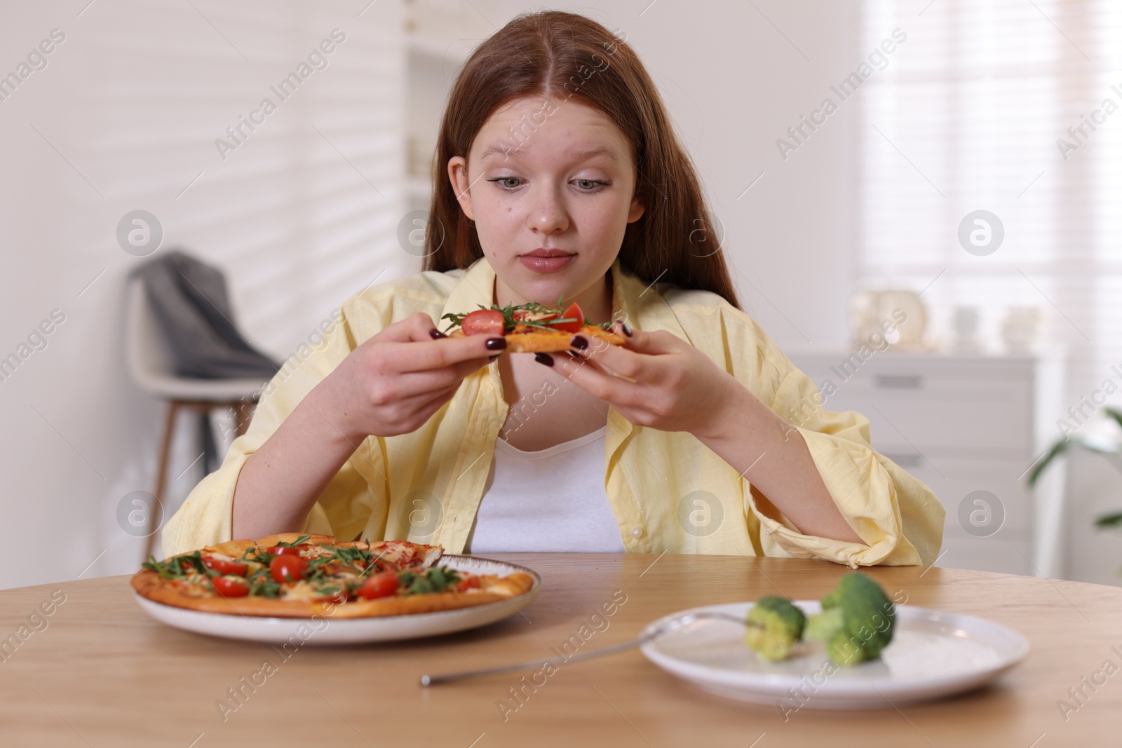 Photo of Sad teenage girl with pizza and broccoli at wooden table indoors. Eating disorder