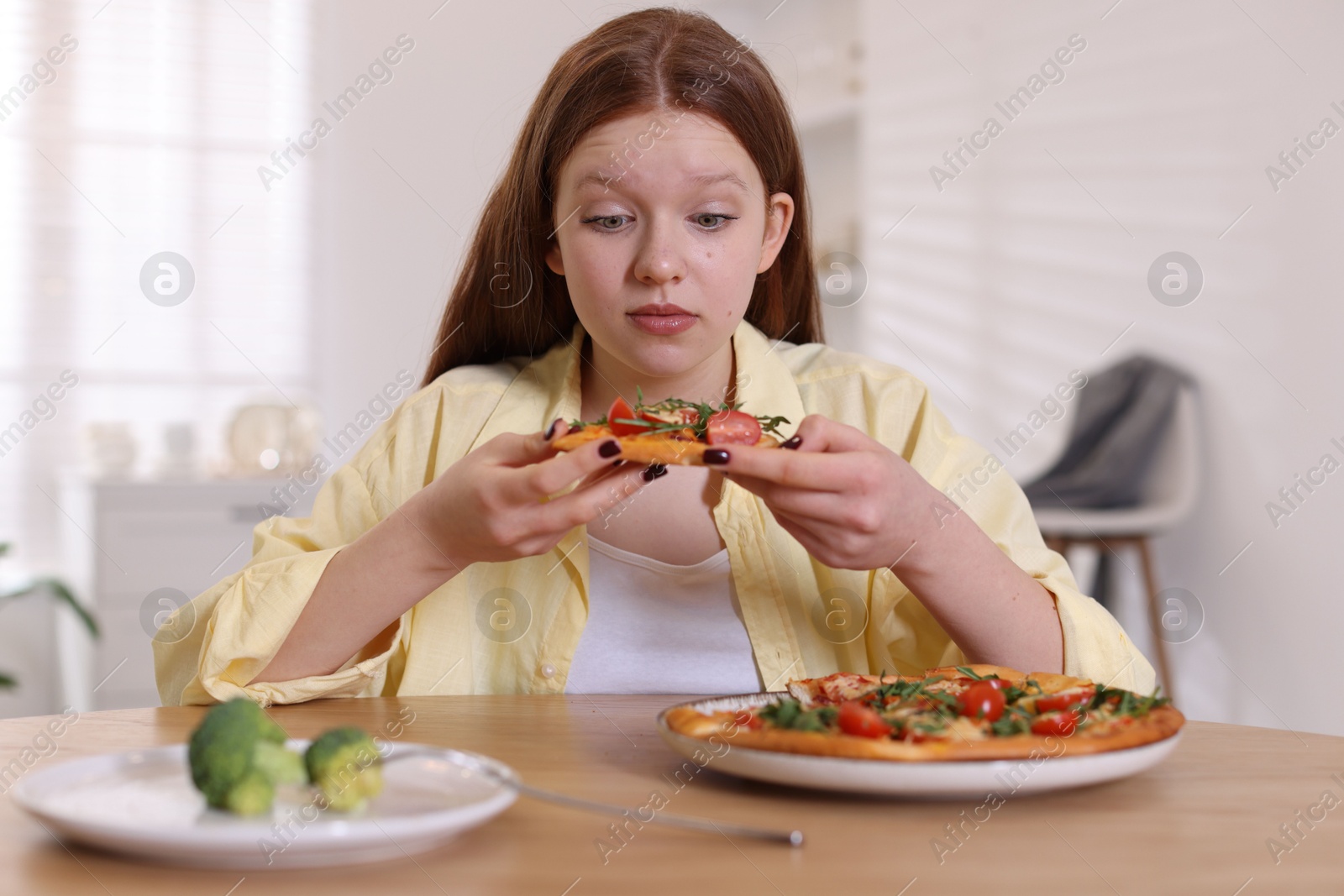 Photo of Sad teenage girl with pizza and broccoli at wooden table indoors. Eating disorder