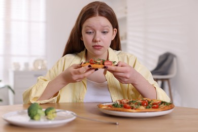 Photo of Sad teenage girl with pizza and broccoli at wooden table indoors. Eating disorder