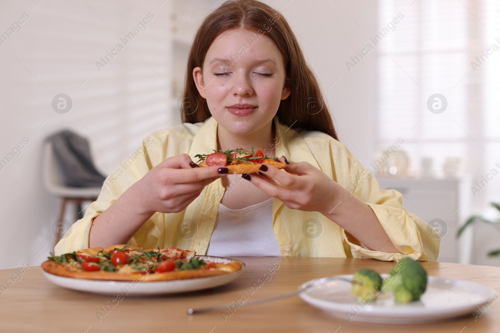 Photo of Sad teenage girl with pizza and broccoli at wooden table indoors. Eating disorder