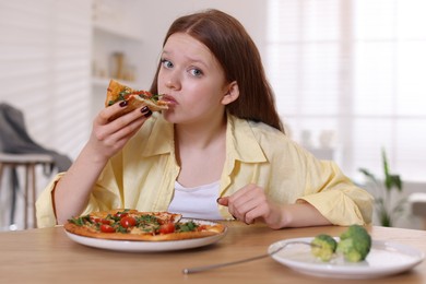 Sad teenage girl with piece of pizza at wooden table indoors. Eating disorder