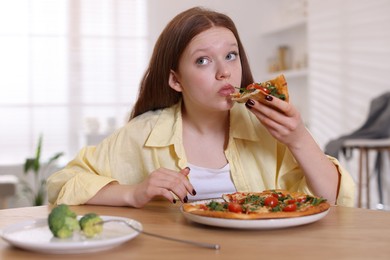 Photo of Sad teenage girl with piece of pizza at wooden table indoors. Eating disorder