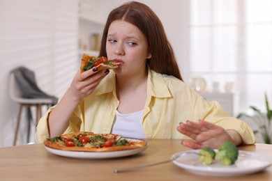 Sad teenage girl with piece of pizza at wooden table indoors. Eating disorder