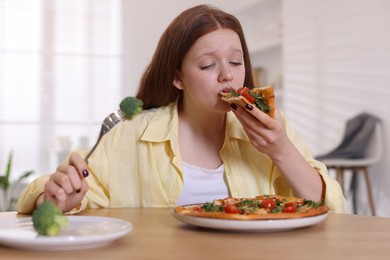 Photo of Sad teenage girl with piece of pizza at wooden table indoors. Eating disorder