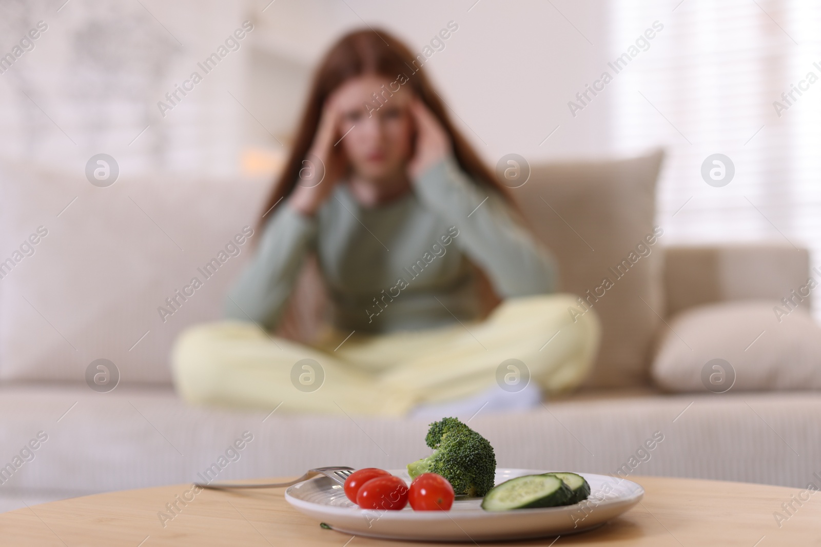 Photo of Sad teenage girl sitting on sofa near table with vegetables at home, selective focus. Eating disorder