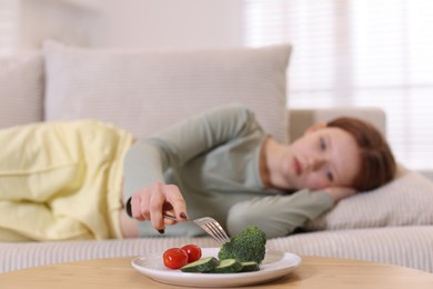 Photo of Sad teenage girl lying on sofa near table with vegetables at home, selective focus. Eating disorder