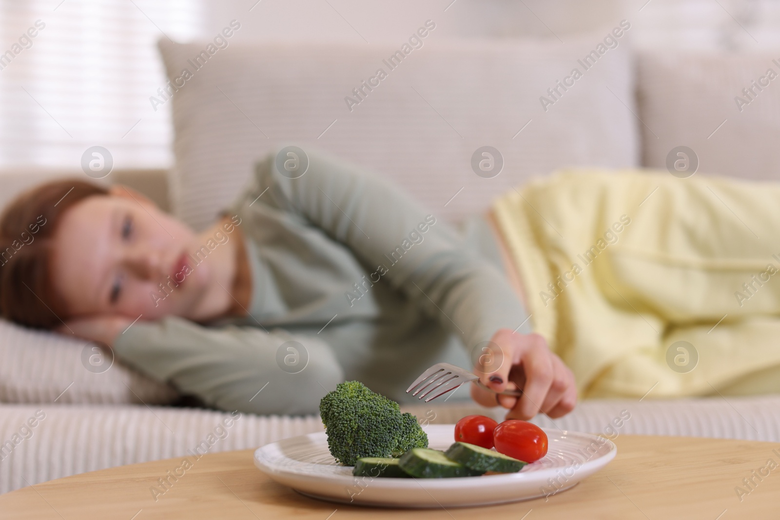 Photo of Sad teenage girl lying on sofa near table with vegetables at home, selective focus. Eating disorder