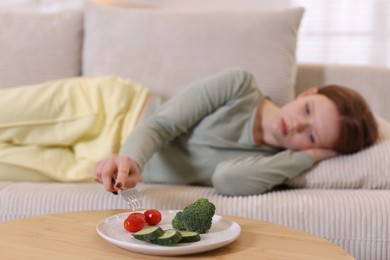 Photo of Sad teenage girl lying on sofa near table with vegetables at home, selective focus. Eating disorder