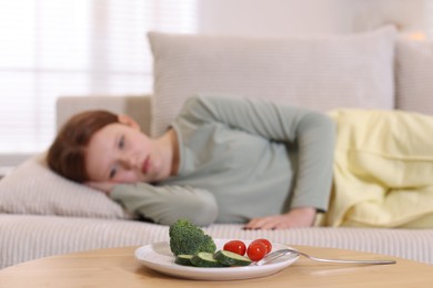 Photo of Sad teenage girl lying on sofa near table with vegetables at home, selective focus. Eating disorder