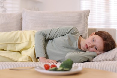 Photo of Sad teenage girl lying on sofa near table with vegetables at home. Eating disorder