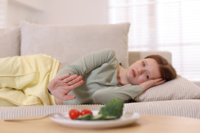 Photo of Sad teenage girl lying on sofa and making stop gesture near table with vegetables at home, selective focus. Eating disorder