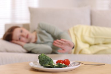 Photo of Sad teenage girl lying on sofa and making stop gesture near table with vegetables at home, selective focus. Eating disorder