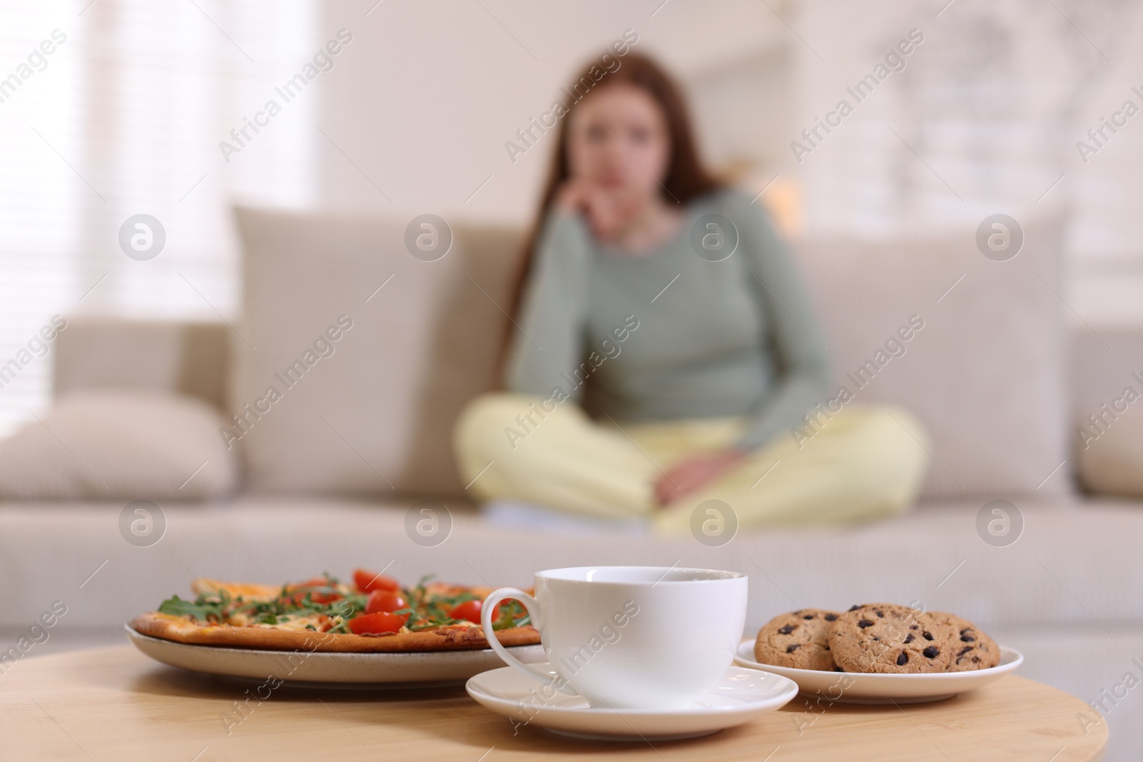 Photo of Sad teenage girl sitting on sofa near table with pizza, coffee and cookies at home, selective focus. Eating disorder