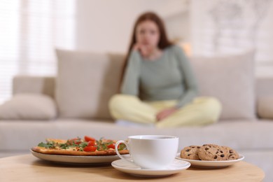 Sad teenage girl sitting on sofa near table with pizza, coffee and cookies at home, selective focus. Eating disorder