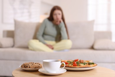 Photo of Sad teenage girl sitting on sofa near table with pizza, coffee and cookies at home, selective focus. Eating disorder