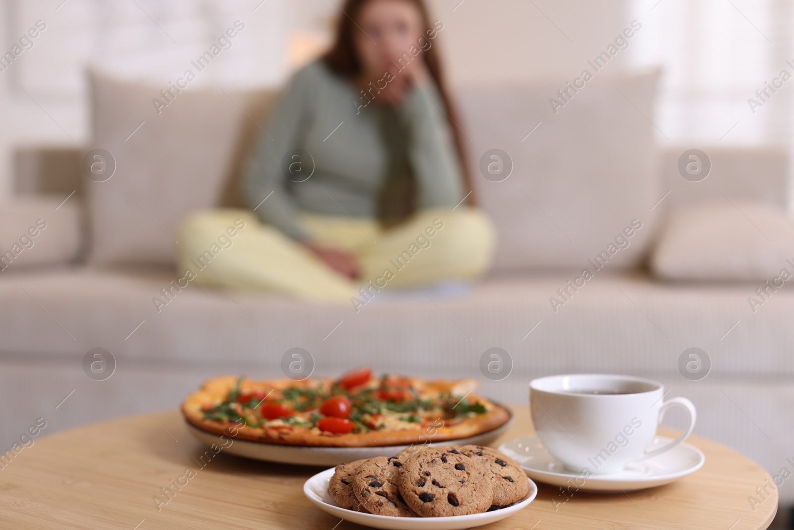 Photo of Sad teenage girl sitting on sofa near table with pizza, coffee and cookies at home, selective focus. Eating disorder