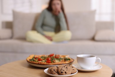 Sad teenage girl sitting on sofa near table with pizza, coffee and cookies at home, selective focus. Eating disorder