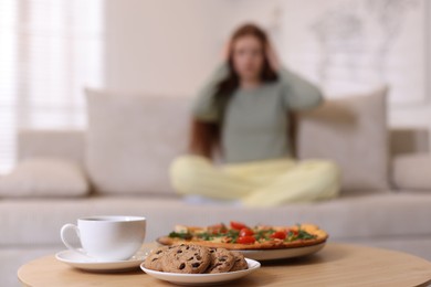 Photo of Sad teenage girl sitting on sofa near table with pizza, coffee and cookies at home, selective focus. Eating disorder