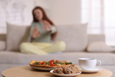 Photo of Sad teenage girl sitting on sofa and making stop gesture near table with pizza, coffee and cookies at home, selective focus. Eating disorder