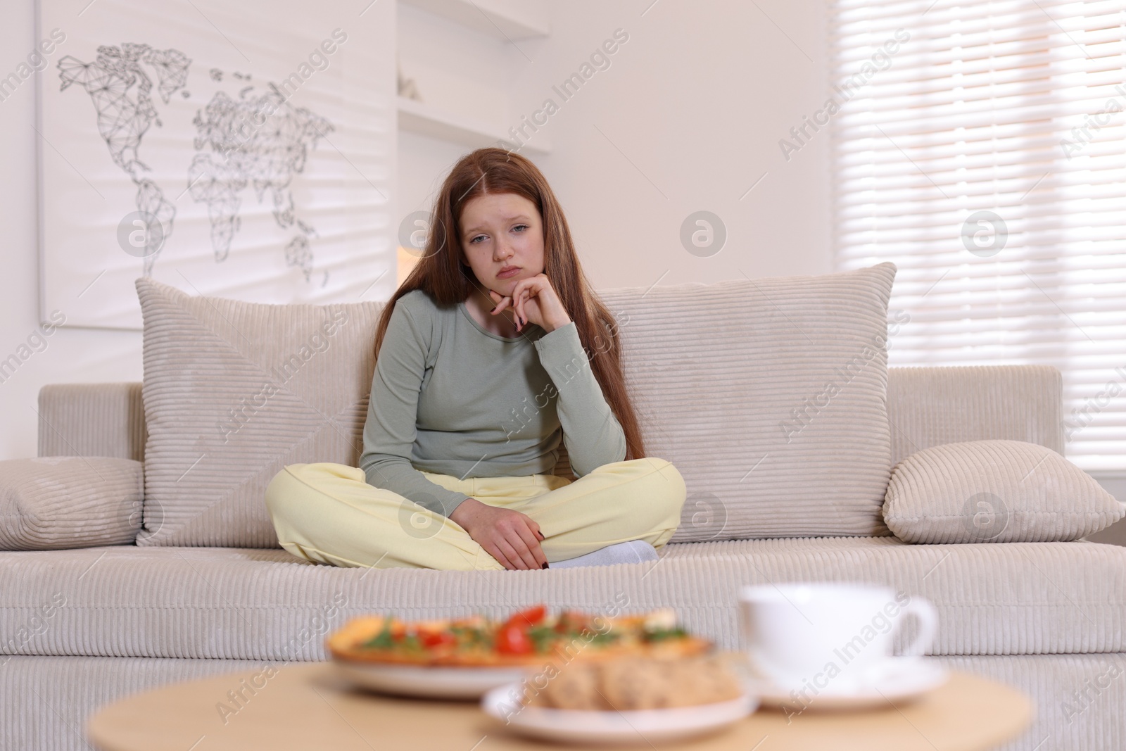 Photo of Sad teenage girl sitting on sofa near table with pizza, coffee and cookies at home. Eating disorder