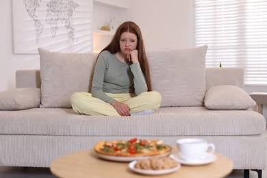 Photo of Sad teenage girl sitting on sofa near table with pizza, coffee and cookies at home. Eating disorder