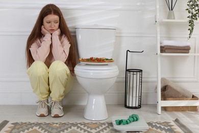 Sad teenage girl sitting near toilet bowl, pizza, scales and measuring tape in bathroom. Bulimia