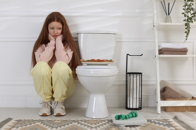 Photo of Sad teenage girl sitting near toilet bowl, pizza, scales and measuring tape in bathroom. Bulimia