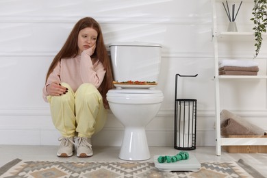 Photo of Sad teenage girl sitting near toilet bowl, pizza, scales and measuring tape in bathroom. Bulimia