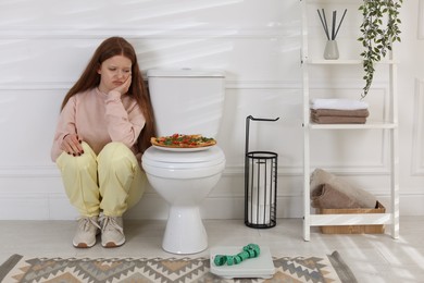 Sad teenage girl sitting near toilet bowl, pizza, scales and measuring tape in bathroom. Bulimia