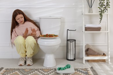 Photo of Sad teenage girl sitting near toilet bowl, pizza, scales and measuring tape in bathroom. Bulimia