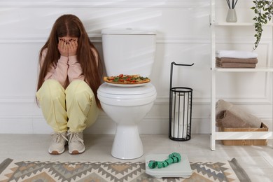 Photo of Sad teenage girl sitting near toilet bowl, pizza, scales and measuring tape in bathroom. Bulimia