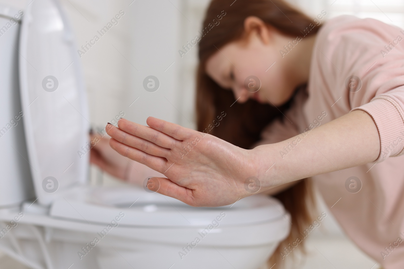 Photo of Teenage girl suffering from nausea over toilet bowl and making stop gesture in bathroom, selective focus. Bulimia