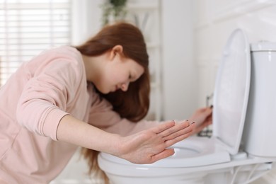 Photo of Teenage girl suffering from nausea over toilet bowl and making stop gesture in bathroom, selective focus. Bulimia