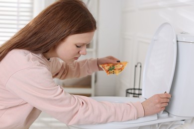 Photo of Sad teenage girl with pizza near toilet bowl in bathroom. Bulimia