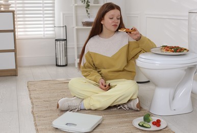 Photo of Sad teenage girl with pizza near toilet bowl in bathroom. Bulimia