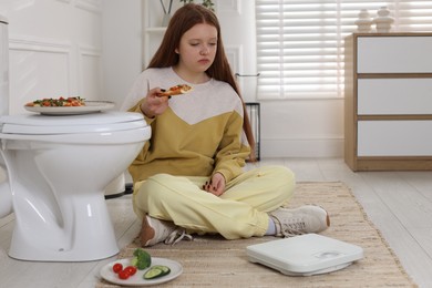 Photo of Sad teenage girl with pizza near toilet bowl in bathroom. Bulimia