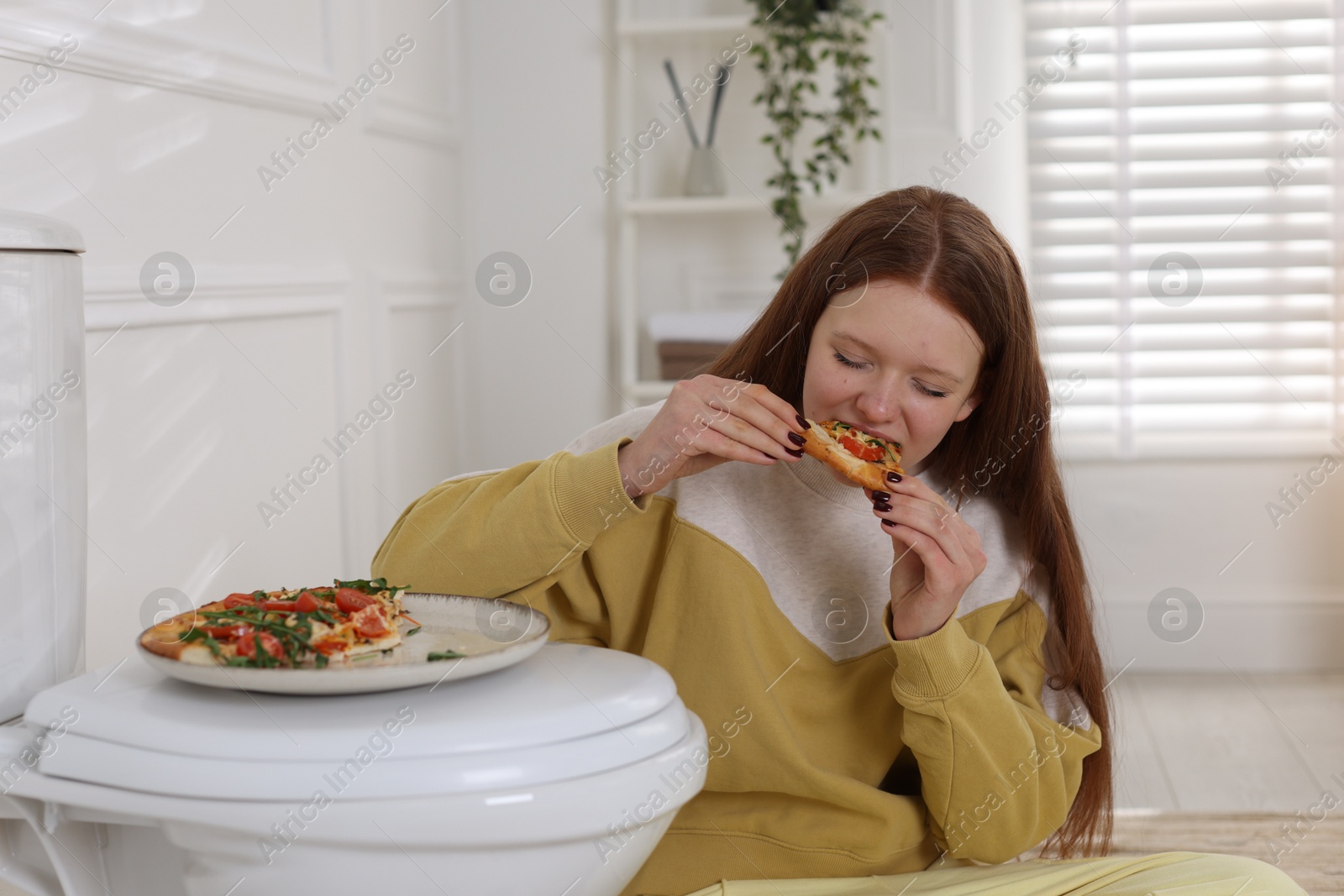 Photo of Teenage girl eating pizza near toilet bowl in bathroom. Bulimia