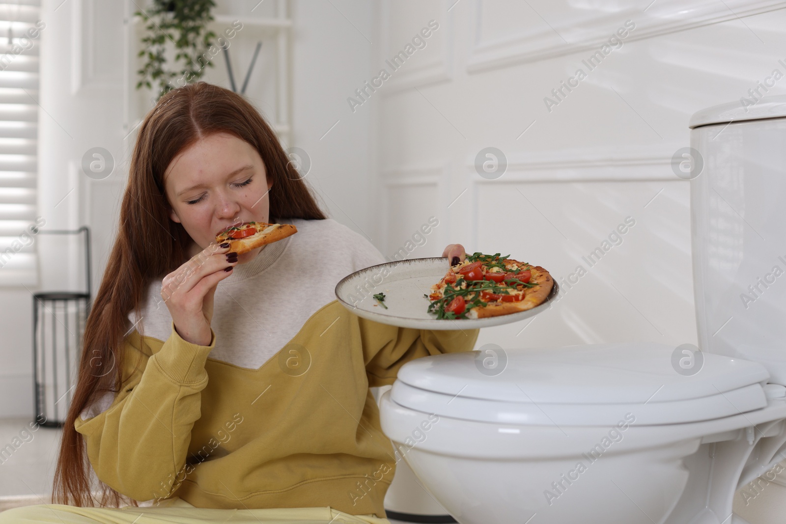 Photo of Teenage girl eating pizza near toilet bowl in bathroom. Bulimia