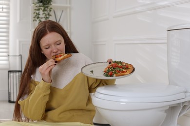 Teenage girl eating pizza near toilet bowl in bathroom. Bulimia