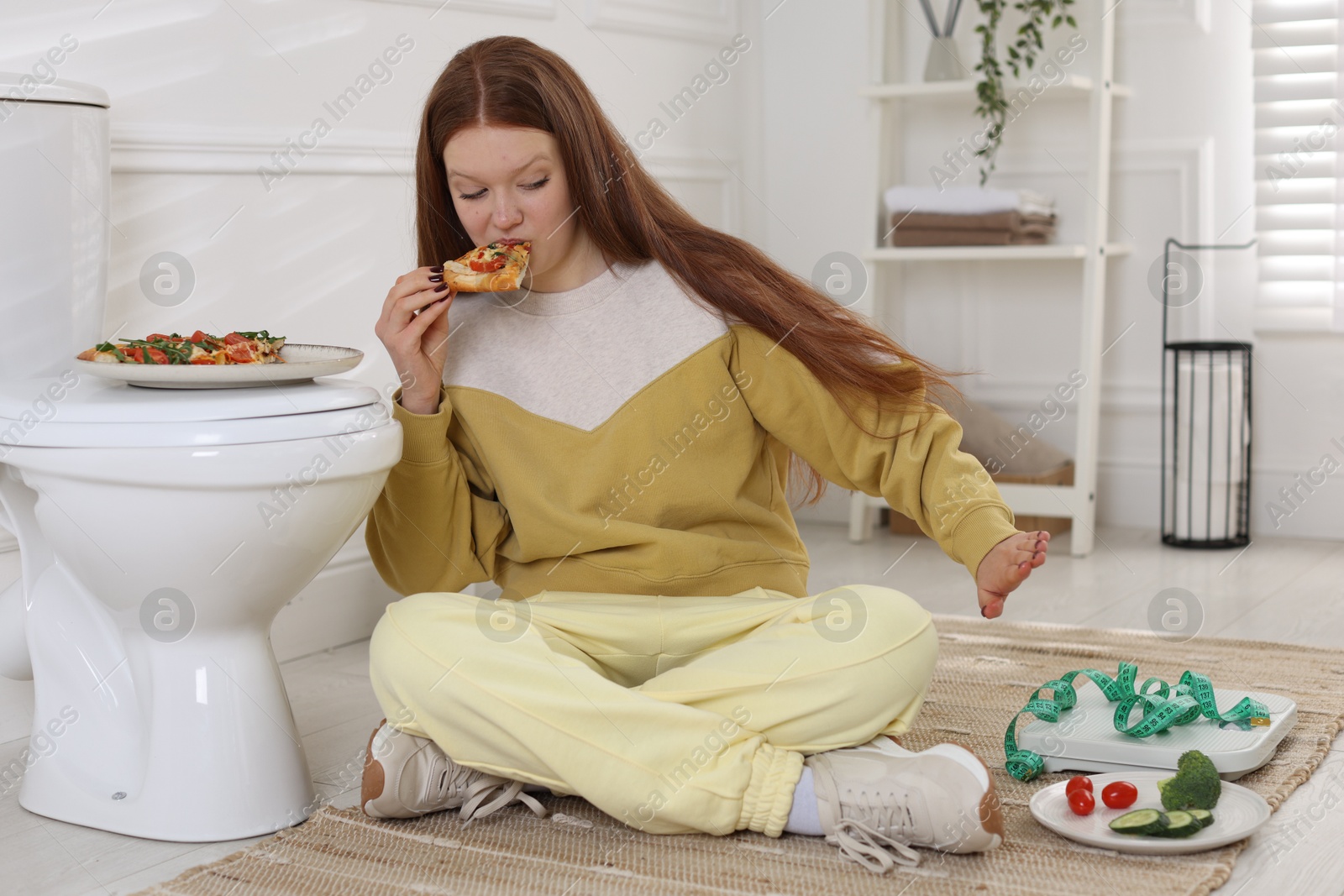 Photo of Teenage girl eating pizza near toilet bowl in bathroom. Bulimia