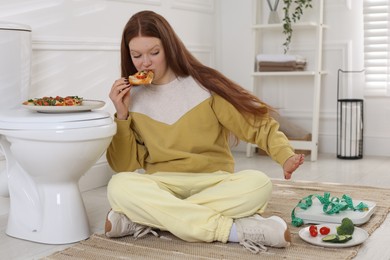 Teenage girl eating pizza near toilet bowl in bathroom. Bulimia