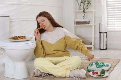 Photo of Teenage girl eating pizza near toilet bowl in bathroom. Bulimia