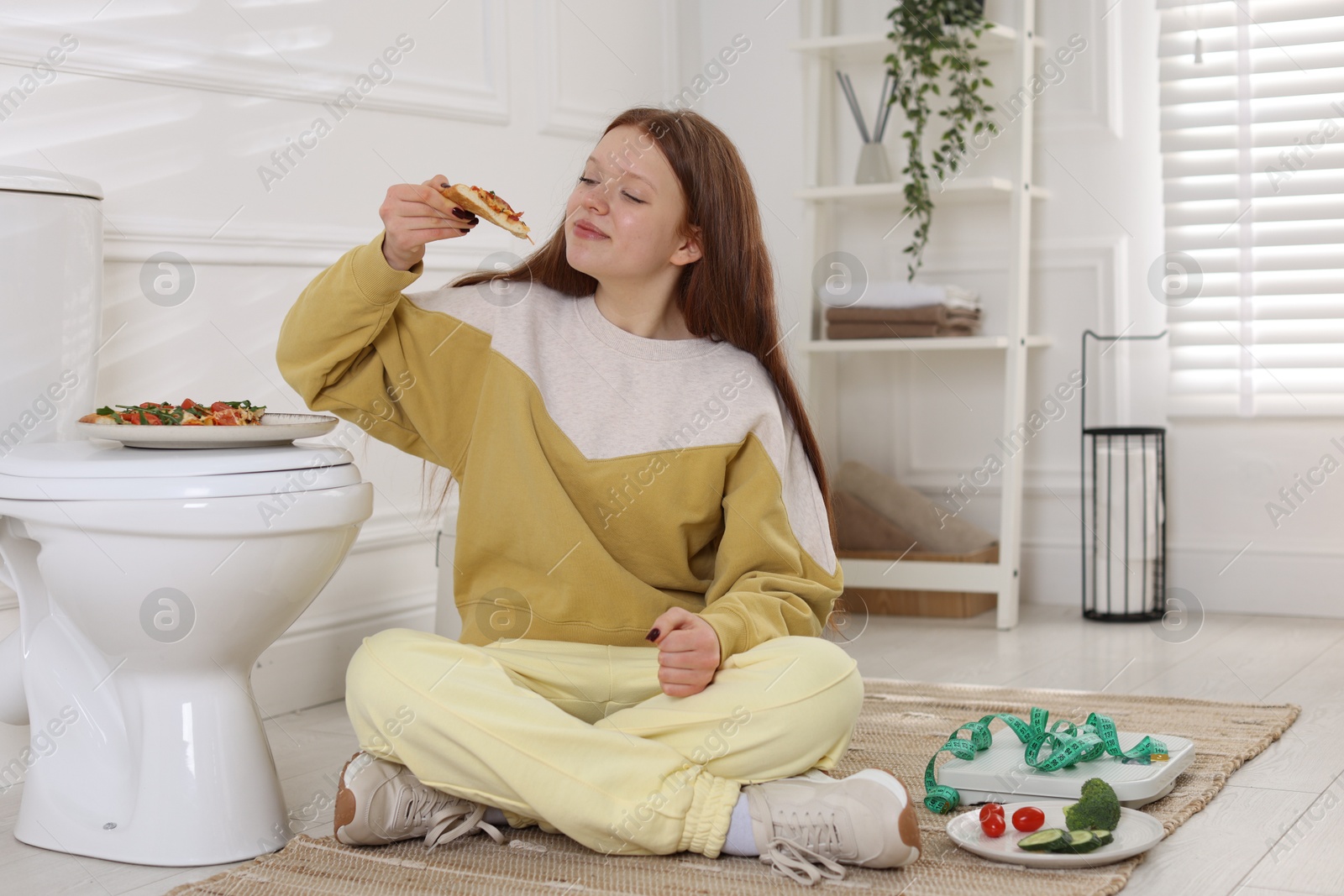 Photo of Teenage girl eating pizza near toilet bowl in bathroom. Bulimia