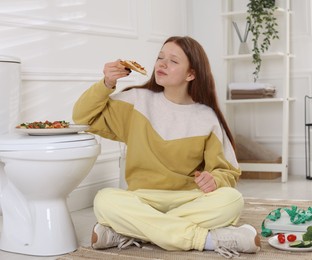 Teenage girl eating pizza near toilet bowl in bathroom. Bulimia