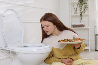 Photo of Sad teenage girl with pizza near toilet bowl in bathroom. Bulimia